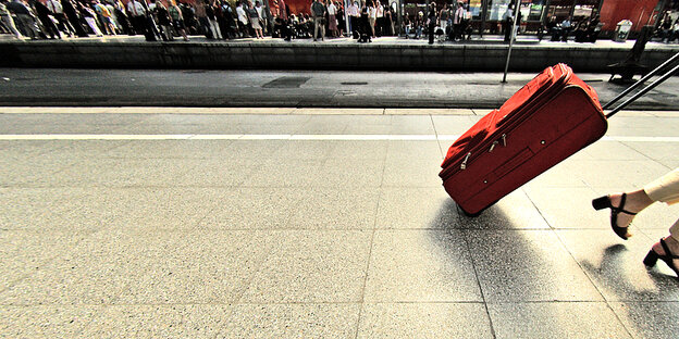 Auf einem Bahnsteig: Frau läuft mit einem roten Rollkoffer aus dem Bild, es sind nur noch ihre Schuhe und Hosenbeine zu sehen