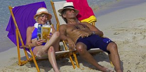 Eine Frau und ein Mann sitzen nebeneinander am Strand, sie liest, er döst