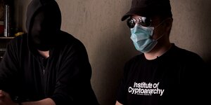 Zwei Menschen sitzen nebeneinander, einem hängt ein schwarzes Tuch über dem Kopf, der andere trägt Kappe, Sonnenbrille, Mundschutz und ein T-Shirt, auf dem „Institute of Cryptoanarchy“ steht