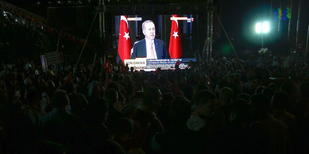 Eine Messenmasse blickt im Dunkeln auf einen Bildschirm, der Recep Tayyip Erdoğan zeigt