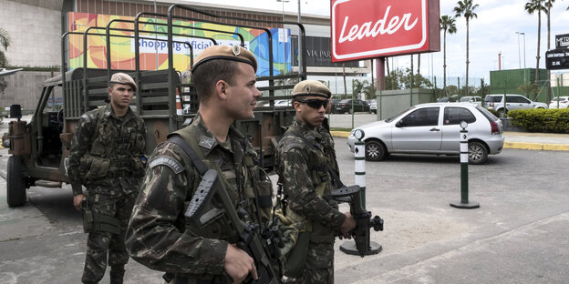 Soldaten mit Gewehren stehen vor einem Gebäude, neben dem eine Werbetafel mit der Aufschrift „Leader“ steht