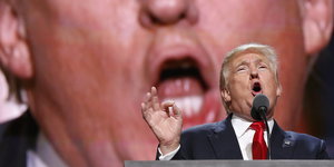 Donald Trump mit aufgerissenem Mund vor einer Leinwand, die sein Gesicht noch einmal zeigt