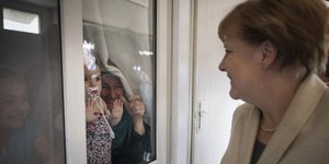 links Kanzlerin Merkel, lachend, rechts hinter einer Fensterscheibe Menschen, die ihr winken