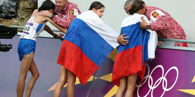 Drei Leichtathleten stehen an der Bande, zwei von ihnen haben sich die russische Nationalflagge übergehängt