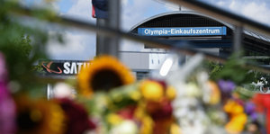 Auf einem Schild steht Olympia-Einkaufszentrum. Im Vordergrund sind verschwommen Blumen zu erkennen