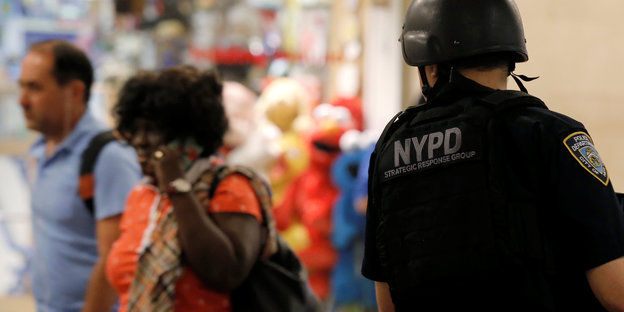 Ein Polizist von hinten, auf seinem Hemd steht "NYPD", er beobachtet zwei Passanten