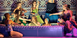 Ein Mann sitzt in einer Art Badewanne, umgeben von Frauen in Haremskleidung – es handelt sich um eine Theaterszene
