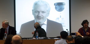 Julian Assange auf einer Leinwand, im Vordergrund sitzen Menschen