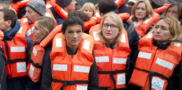 Eine Frau steht zwischen Menschen mit Rettungswesten. Es ist Sahra Wagenknecht. Ein Schild mit dem Schriftzug „Die Linke“ verdeckt ihren Mund