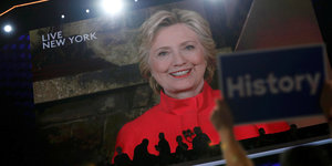Hillary Clinton in einer Videoübertragung, im Vordergrund hält jemand ein Schild, auf dem „History“ steht