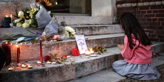 Ein Mädchen mit gefalteten Händen kniet vor Stufen, auf denen Kerzen und Blumen liegen