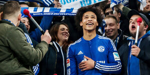 Der Fußballer Leroy Sané steht im Trikot von Schalke 04 zwischen Fans und hält die Hand auf den Bauch