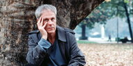 Der Schriftsteller Dennis Cooper sitzt vor einem Baum und stützt seinen Kopf in seine Hand