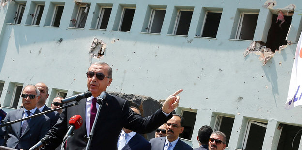 ein Mann in Anzug mit Sonnenbrille (es ist der türkische Präsident Erdogan) vor einem Haus mit Einschusslöchern