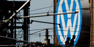 Das VW-Logo an einer Hauswand. Davor diverse Leitungen