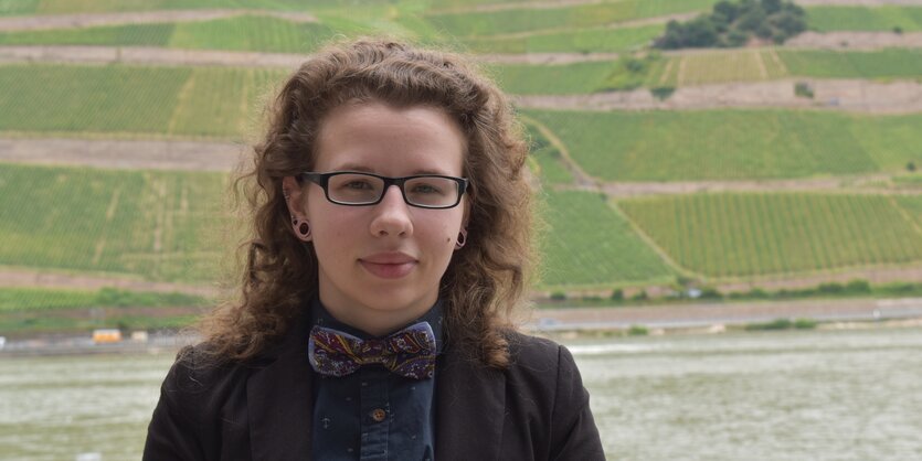 Eine junge Frau mit Locken und Brille steht vor einer grünen Hügellandschaft.