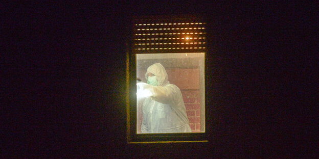 Das Bild zeigt ein Fenster während der Nacht. Im Lichtschein ist eine Person in einem weißen Ganzkörperanzug zu sehen
