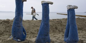 Am Meeresstrand: drei Unterleiber, gekleidet in Jeans, ragen aus dem Sand heraus