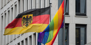 eine Deutschlandflagge und eine Regenbogenflagge wehen im Wind vor einem Gebäude
