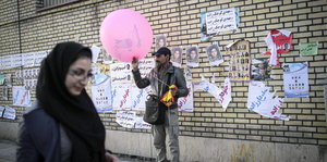 Ein Mann hält einen rosafarbenen Luftballon vor einer Wand, die mit Wahlpostern beklebt ist, im Vordergrund läuft eine Frau vorbei