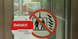 ein „Defekt“-Aufkleber neben einem „Fahrstuhl-im-Brandfall-nicht-benutzen“-Aufkleber