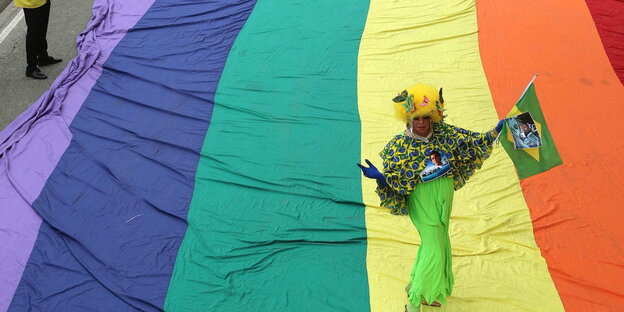 Auf einem Teppich in Regenbogenfarben tanzt eine verkleidete Person mit einer brasilianischen Flagge in der Hand