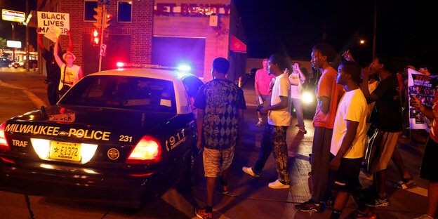 Es ist Nacht, links im Bild ein Polizeiauto und ein Mensch, der ein Schild hochhält, rechts neben dem Polizeiauto mehrere Menschen