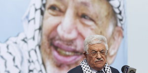 Im Hintergrund ein großes Foto eines Mannes mit Palästinensertuch um den Kopf, im Vordergrund ein älterer Mann mit weiß-grauen Haaren und Brille