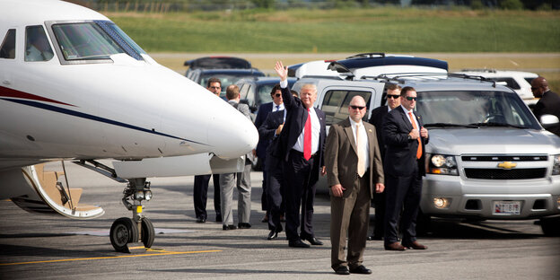Trump steht neben Begleitern auf einem Flugfeld und winkt