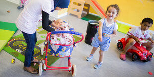 Drei Kinder, eins beugt sich über einen Kinderwagen mit Puppe, ein anderes sitzt auf einem Bobbycar