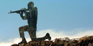 Eine Soldatenfigur mit Gewehr kniet auf einer Steinmauer mit Maschendrahtzaun