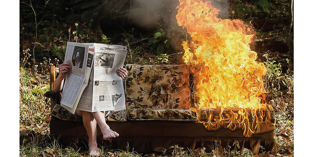 Eine Person sitzt Zeitung lesend auf einem Sofa im Wald, während das Sofa neben ihm anfängt zu brennen.