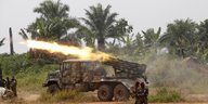 Ein Armeegeschütz feuert Munition ab, ein Feuerstrahl schießt durch die Luft. Dahinter grüne Palmen