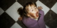 Ein kleines Mädchen sitzt mit weit geöffnetem Mund auf einem schwarz-weiß-gefliesten Boden