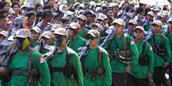 Uniformierte Menschen mit Gesichtsbemalung und Gewehren marschieren im Gleichschritt