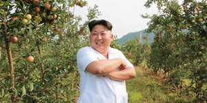 Kim Jong-un steht grinsend und mit verschränkten Armen in einer Apfelplantage