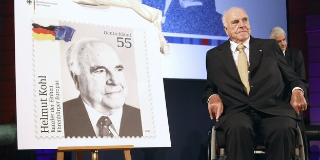 Helmut Kohl sitzt im Rollstuhl neben einer sehr großen Helmut-Kohl-Briefmarke