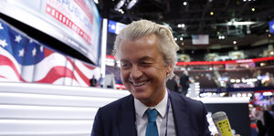 Ein Mann mit grauen Haaren trägt Anzug und Krawatte und lacht. Im Hintergrund US-amerikanische Flaggen-Dekoration
