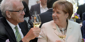 Ein älterer Mann mit grauen Haaren und eine Frau mit kurzen rot-blonden Haaren halten ihre Weingläser zum Anstoßen hoch