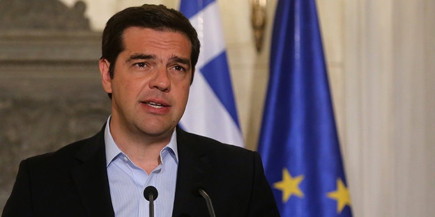 Ein Mann mit dunklen Haaren spricht in ein Mikrofon, hinter ihm die griechische und die europäische Flagge