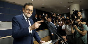 Rajoy lächelt und zeigt auf etwas bei einer Pressekonferenz, im Hintergrund viele FotografInnen