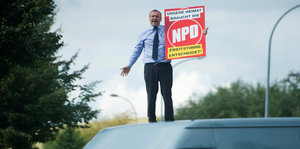 Ein Mann in Anzug steht auf einem Autodach und hält ein Wahlplakat der NPD in der Hand