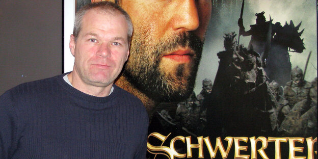Ein Mann mit kurzen grauen Haaren steht vor einem großen Filmplakat