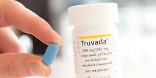 Zwei Finger halten eine kleine blaue Pille, dahinter die Pillendose mit Aufschrift „Truvada“