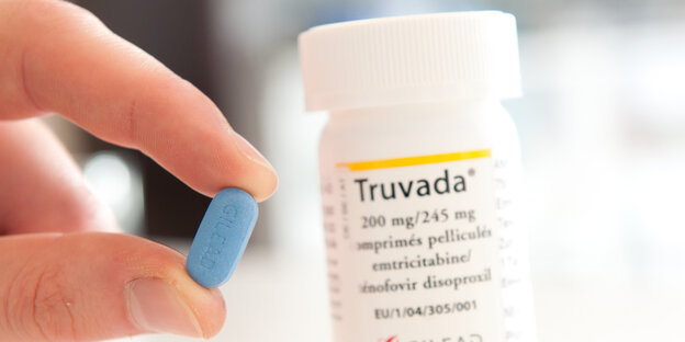 Zwei Finger halten eine kleine blaue Pille, dahinter die Pillendose mit Aufschrift „Truvada“