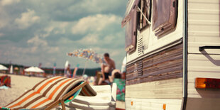 Ein Wohnwagen steht neben einer Liege am Strand, im Hintergrund stehen Menschen