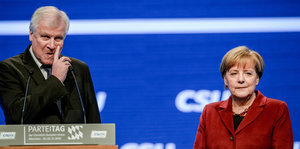 Seehofer steht am Rednerpult und kratzt sich im Gesicht, Merkel steht daneben