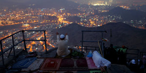 Auf einem Berg mit Blick auf eine beleuchtete Stadt betet ein Mann auf einem ausgebreiteten Teppich