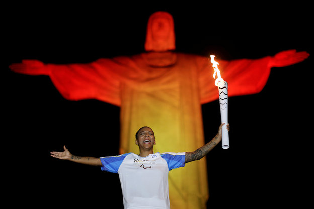 Vor der gelb-rot beleuchteten Christus-Statue von Rio steht eine brasilianische Sportlerin mit ausgebreiteten Armen, in der einen Hand eine Fackel
