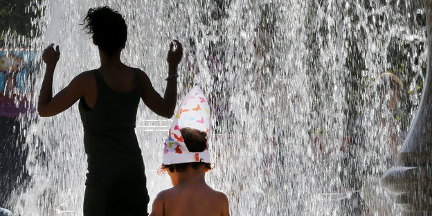Eine Frau an einem Springbrunnen dreht ihrem Kind den Rücken zu. Das Kind sitzt am Springbrunnenrand mit einem Eimer